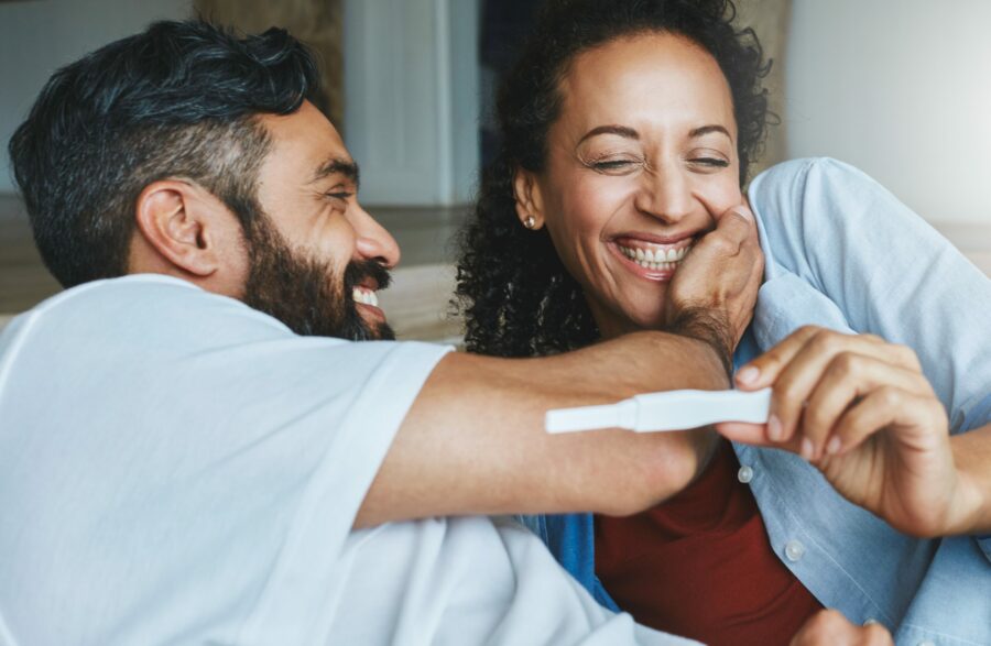 Imagem de homem e mulher indianos sorrindo. A mulher segura e olha um teste de gravidez enquanto o homem toca carinhosamente seu rosto.