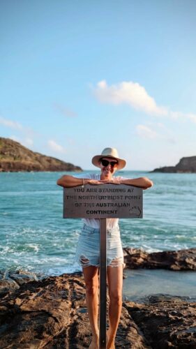 Afbeelding van Lauren die naar de camera lacht, terwijl ze op het strand is. Lauren staat op de rotsen en leunt op een bord met de tekst "Je staat op het meest noordelijke punt van het Australische continent"